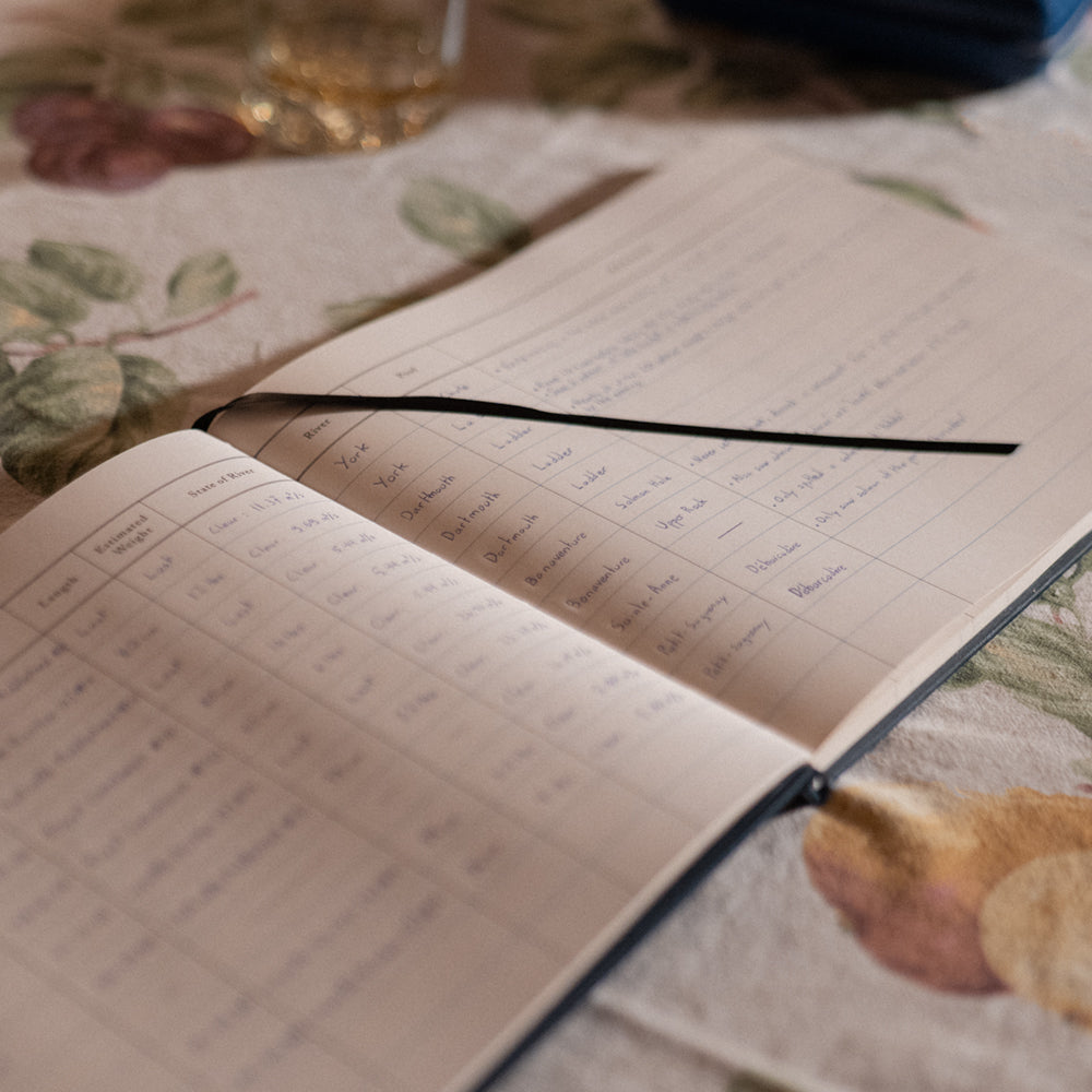Interior pages of the Atlantic Salmon Log showing fields to record fly, river, and conditions laying on a table with a glass of Scotch.