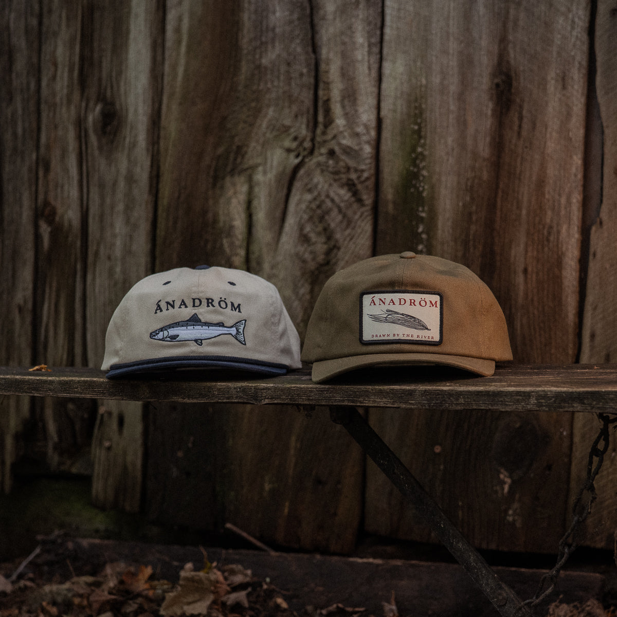 Two vintage fly fishing Ánadröm branded caps on a wooden surface with a rustic background.