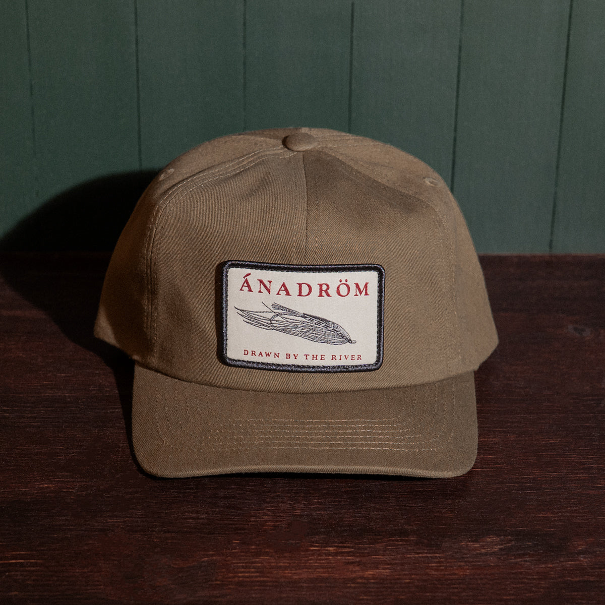 Olive cap with a fly fishing embroidered patch on a wooden surface against a green wall
