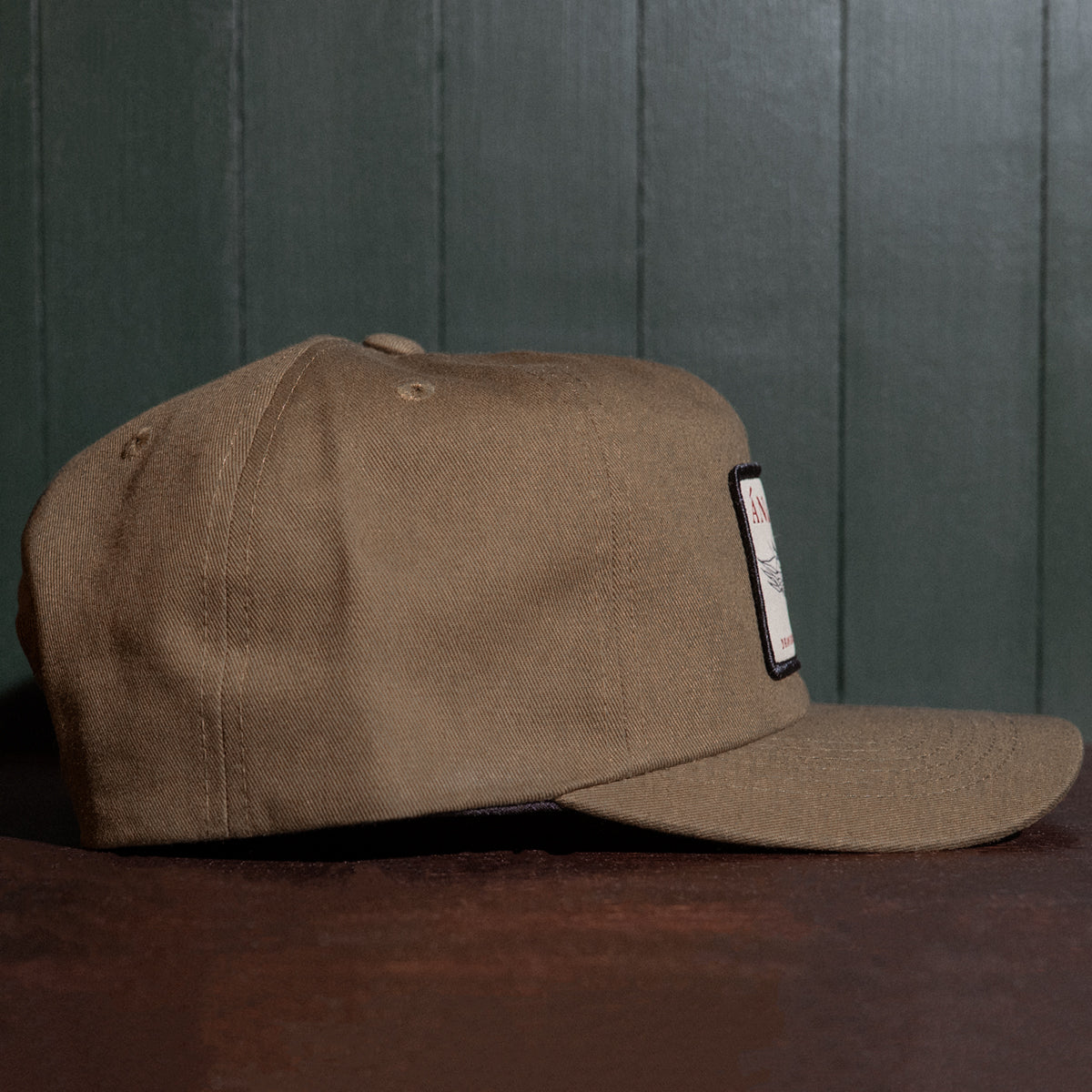 Olive cap from the side with a salmon fly patch on a dark wood surface and green wooden panel background