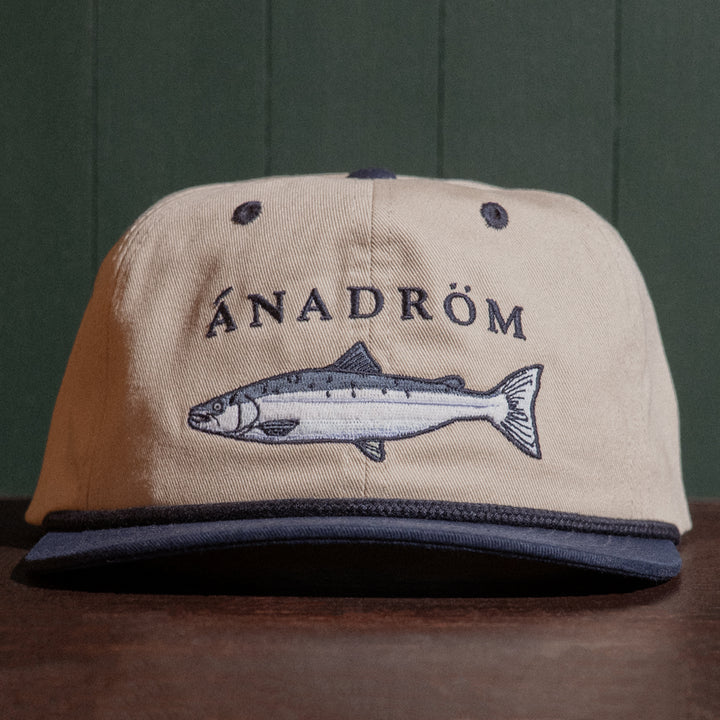 Vintage stone coloured cap with salmon embroidery and 'ÁNADRÖM' text on a wooden table and green background