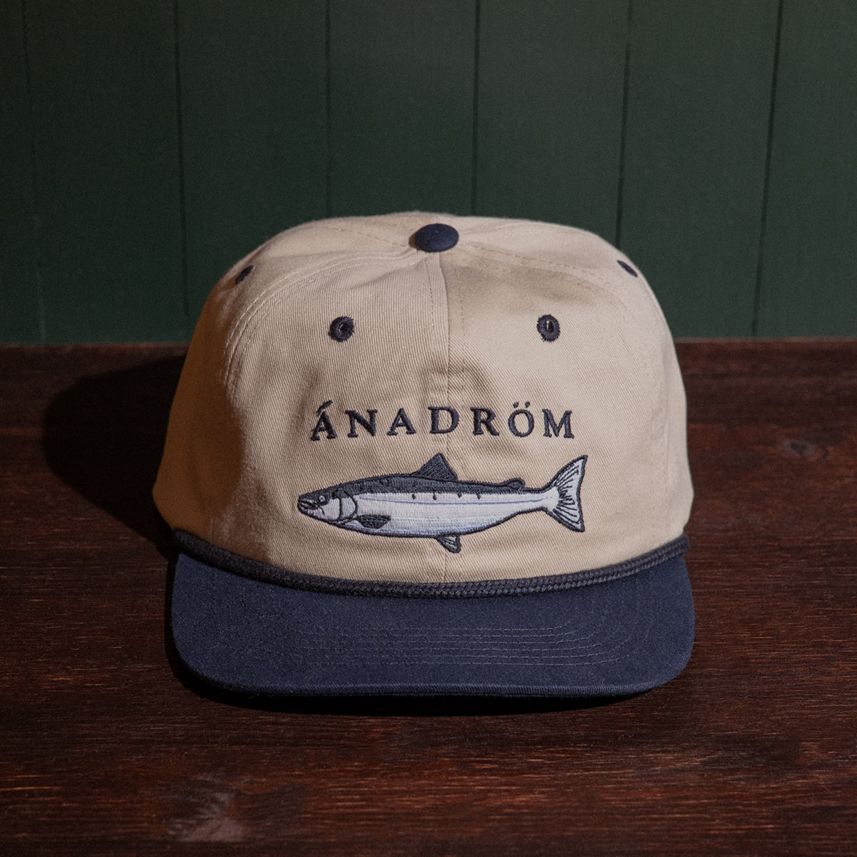 Vintage stone and navy cap with salmon embroidery design and 'Ánadróm' text on a wooden surface.