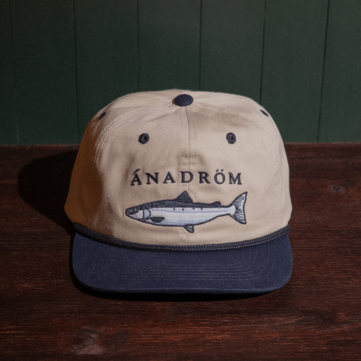 Vintage stone and navy cap with salmon embroidery design and 'Ánadróm' text on a wooden surface.