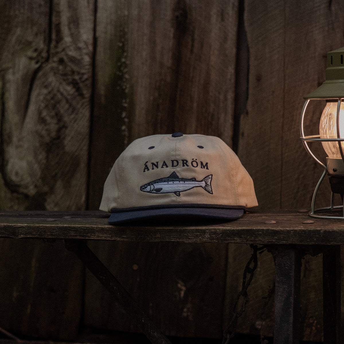 Stone and navy cap with a salmon design and 'ÁNADRÖM' logo on a wooden surface with a rustic background.