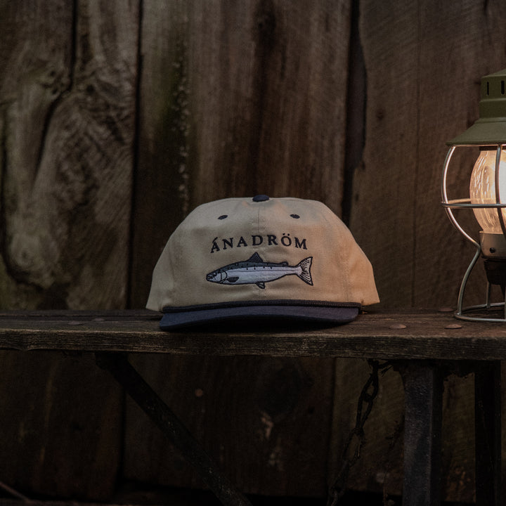 Stone and navy cap with a salmon design and 'ÁNADRÖM' logo on a wooden surface with a rustic background.