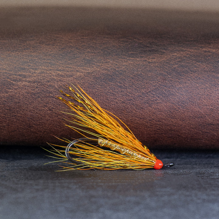 Atlantic salmon fly with yellow, orange, black hair, and a gold body, called the Willie Gun with a leather wallet in the background
